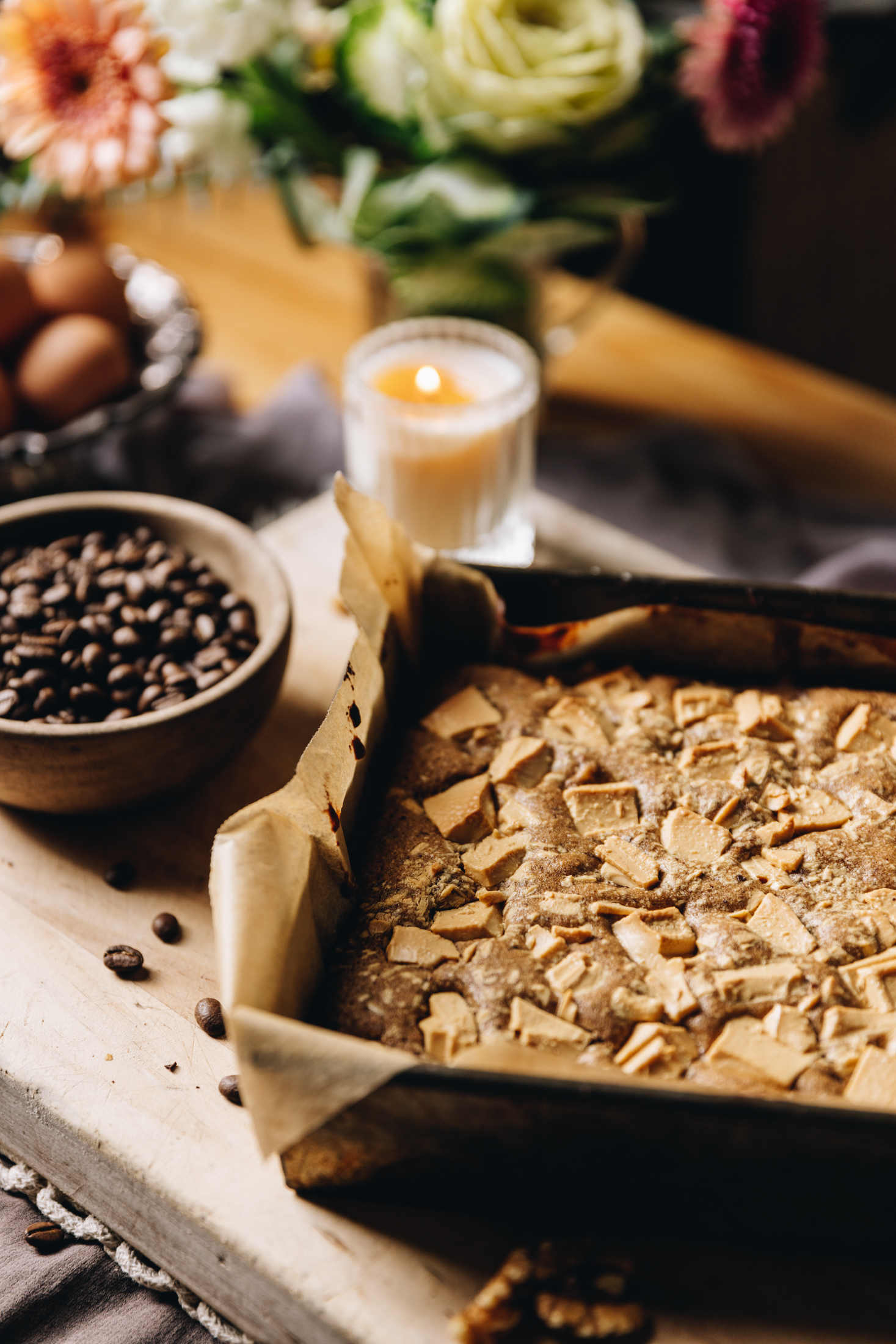 Baked coffee blondie with caramelised white chocolate on top. Coffee beans, a candle and flowers in the background. 