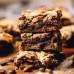Three chocolate coffee blondies with walnuts stacked on a wooden board with gooey dark chocolate. Coffee beans sprinkled on the board.