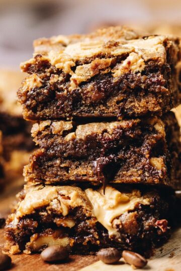 Three chocolate coffee blondies with walnuts stacked on a wooden board with gooey dark chocolate. Coffee beans sprinkled on the board.