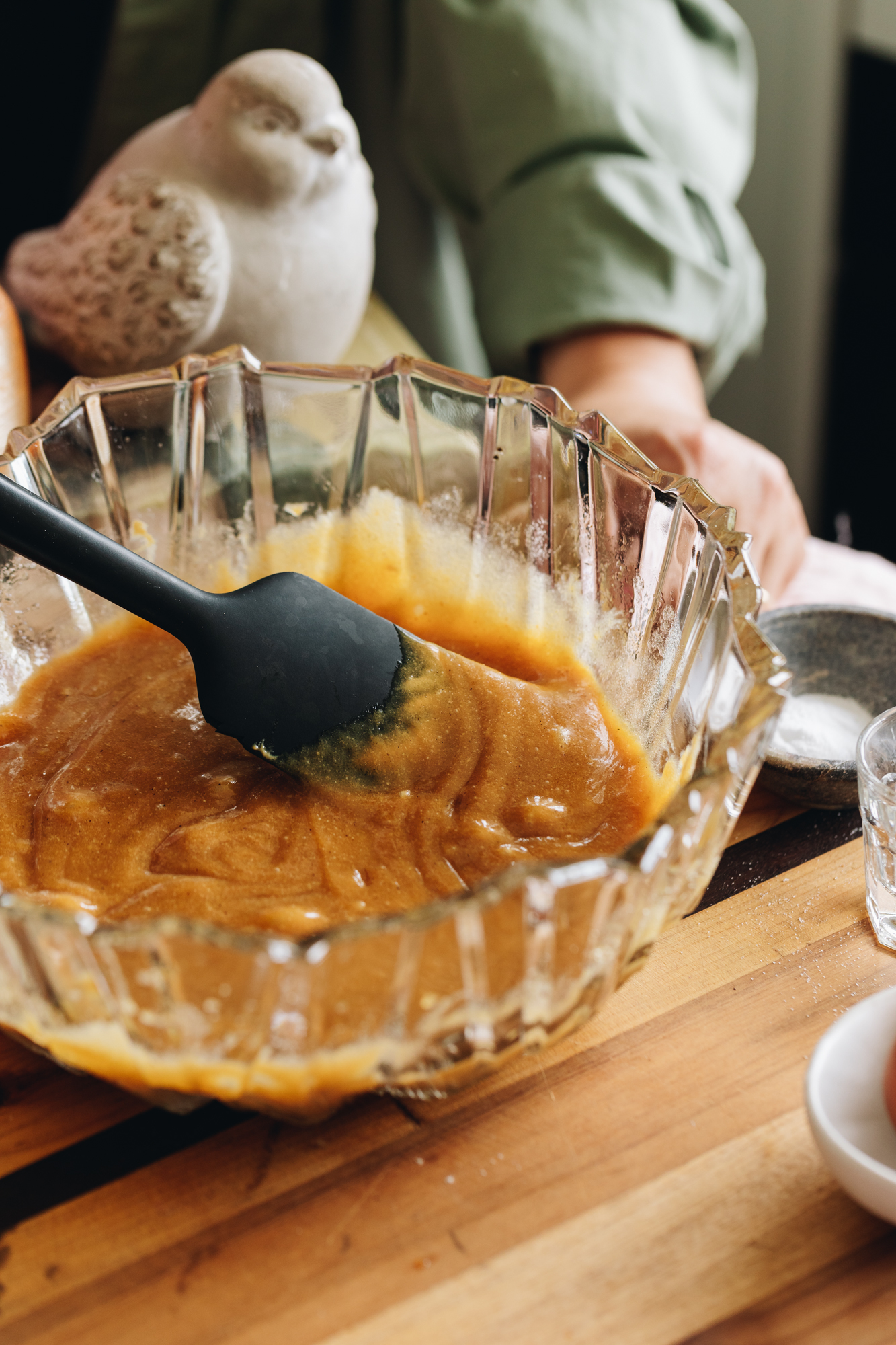 A large glass bowl with butter and eggs mixed together on a wooden table with a concrete bird in the background. 