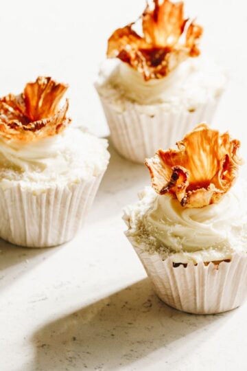 Three decorated hummingbird muffins, topped with cream cheese frosting and dried pineapple flowers.