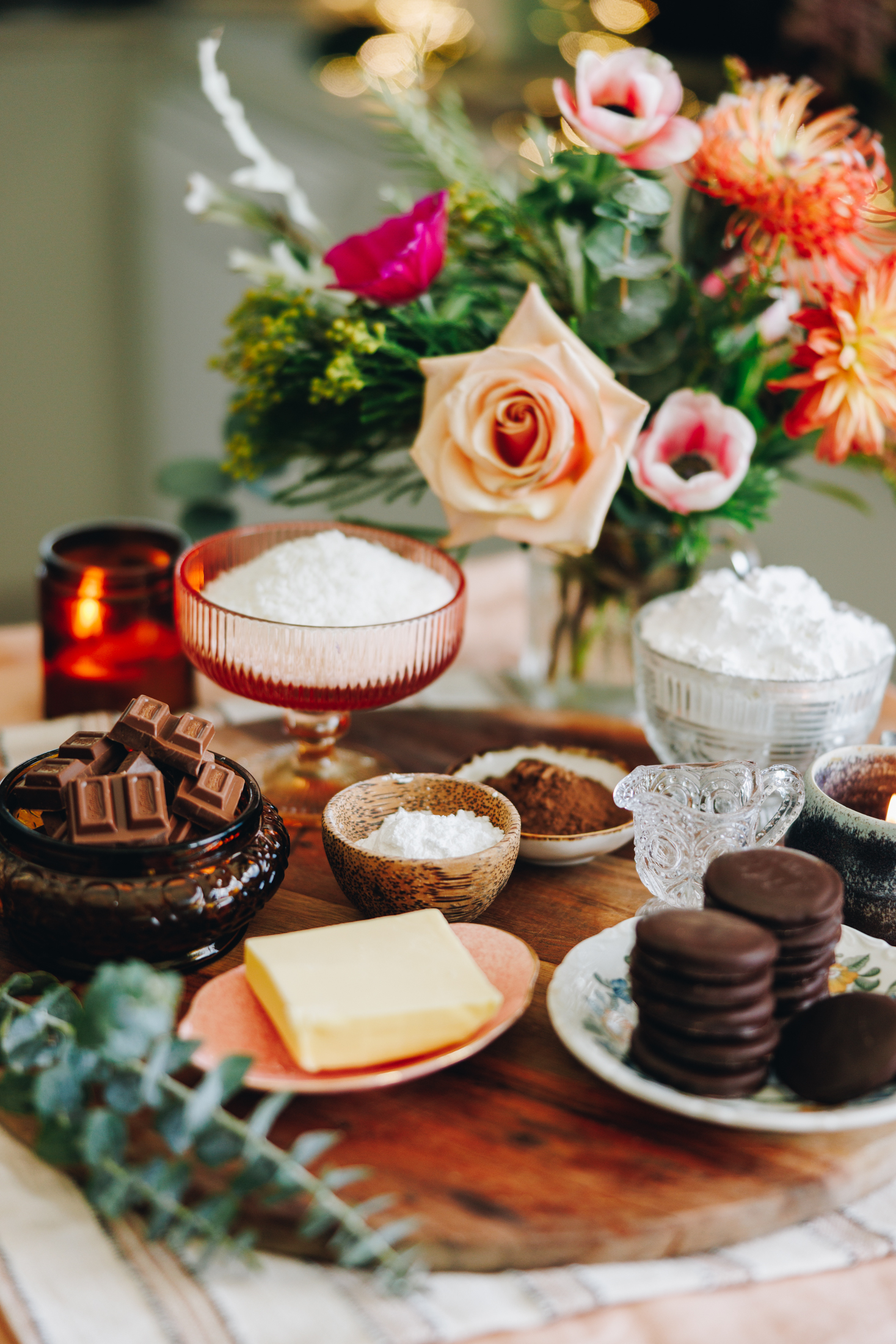 A wooden board with ingredients in vintage bowls. Eucalyptus at the front and flowers in the background with an orange candle.