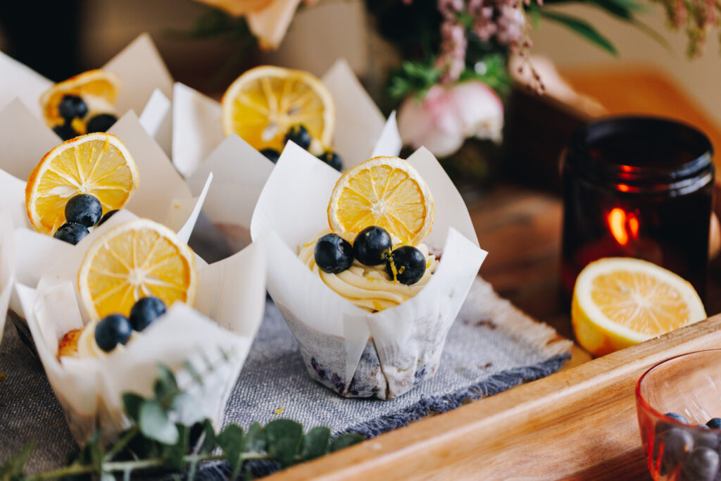 Blueberry and white chocolate muffins decorated with piped cream cheese frosting, dried lemon slices and fresh blueberries on a wooden board with a candle and flowers.